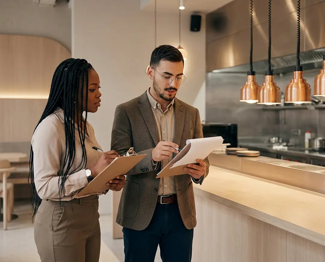 Two people in business attire review documents on clipboards in a modern kitchen setting. They appear focused and engaged in discussion.