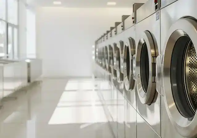 Row of industrial washing machines in a bright, modern laundromat. Sunlight streams through large windows, casting reflections on the glossy floor.