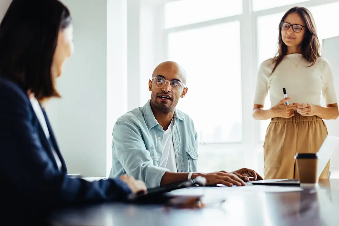 Three people in a bright office. A man in glasses speaks while a woman writes and another listens, creating a collaborative and focused atmosphere.