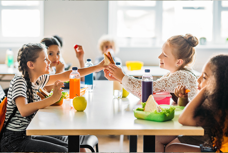 Children sit at a bright cafeteria table, smiling and sharing snacks. Drinks and fruits are visible, creating a joyful and sociable atmosphere.