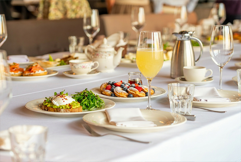 A lavish breakfast table is set, featuring poached egg on avocado toast, berries, pastries, a glass of juice, and tea on fine china. The ambiance is elegant.