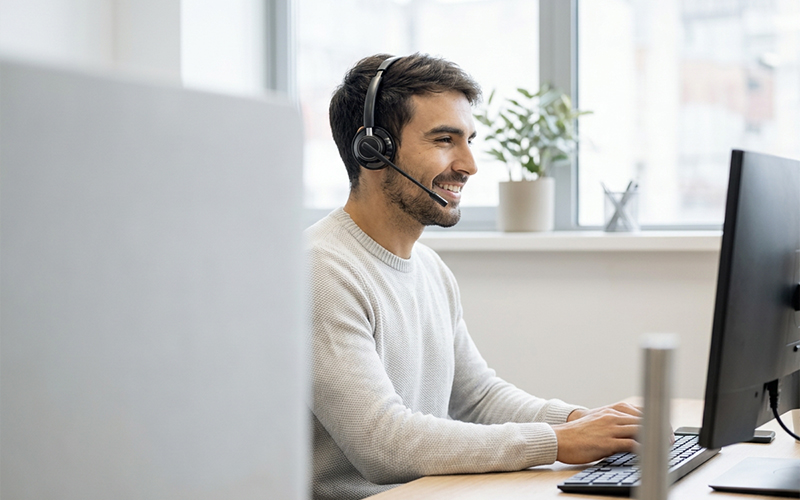 A man wearing a headset smiles while working on a computer in a bright office. A window and a small plant are visible in the background, creating a warm, professional atmosphere.