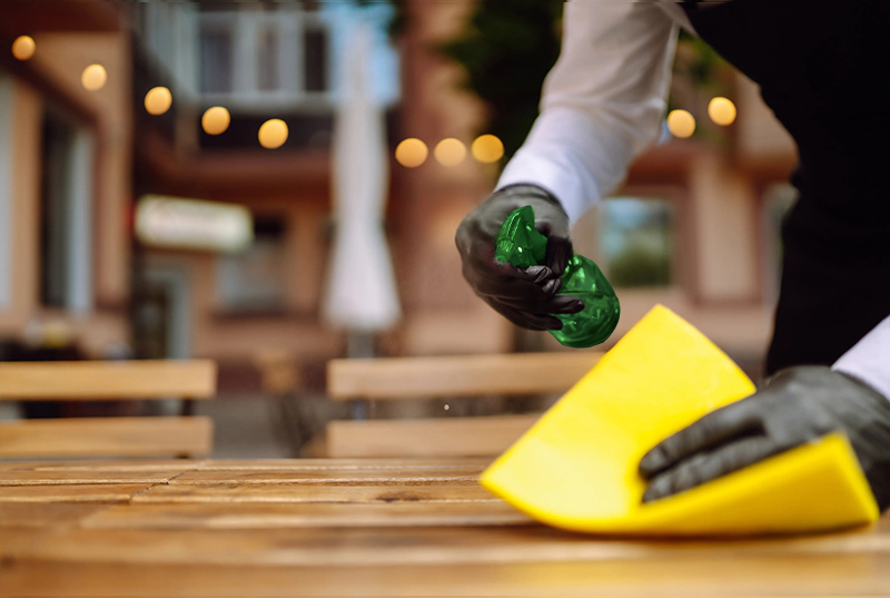 A person wearing gloves cleans a wooden table with a yellow cloth and green spray bottle, set against a blurred outdoor café with string lights.