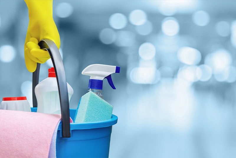 A person wearing yellow gloves holds a blue cleaning bucket filled with spray bottles, sponges, and a pink cloth. The background is blurred and cool-toned.
